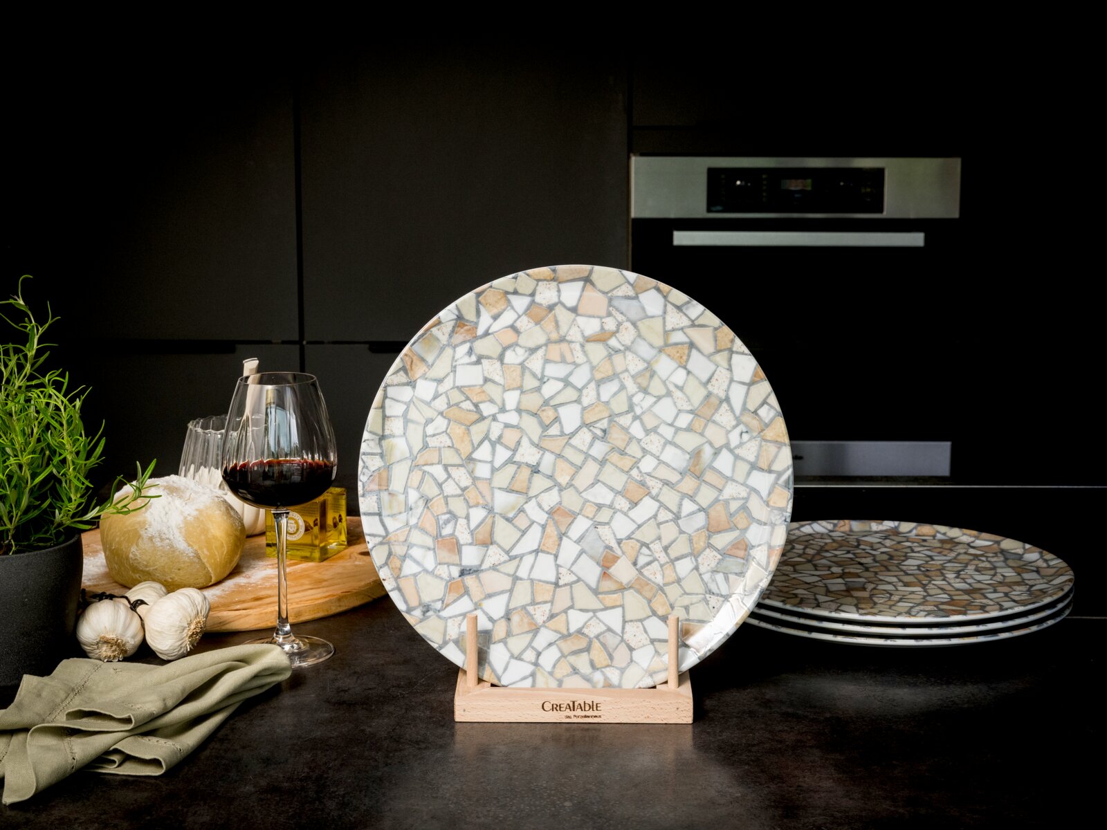 Creatable Pizzateller 4-tlg STONE COLLECTIONF Runder Keramik-Speiseteller mit Mosaik-/Terrazzo-Muster in Beige, Grau und Weiß, frontal aus leicht erhöhter Perspektive auf einem Holzständer in moderner Küche; weitere Teller gestapelt im Hintergrund