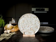 Runder Keramik-Speiseteller mit Mosaik-/Terrazzo-Muster in Beige, Grau und Weiß, frontal aus leicht erhöhter Perspektive auf einem Holzständer in moderner Küche; weitere Teller gestapelt im Hintergrund