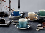 Schräg von vorn fotografiertes Kaffeetassen-Set aus Keramik mit Untertassen in Blau, Beige und Grün auf dunkelgrauem Tisch, dekoriert mit Löffeln, Steinen und getrockneten Zweigen.