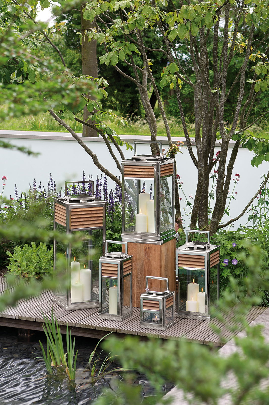 Fink Laterne GENUA Mehrere Laternen aus Glas und Metall mit Holzdetails, auf einer Terrasse im Garten platziert, umgeben von grünen Pflanzen. Perspektive von vorne.