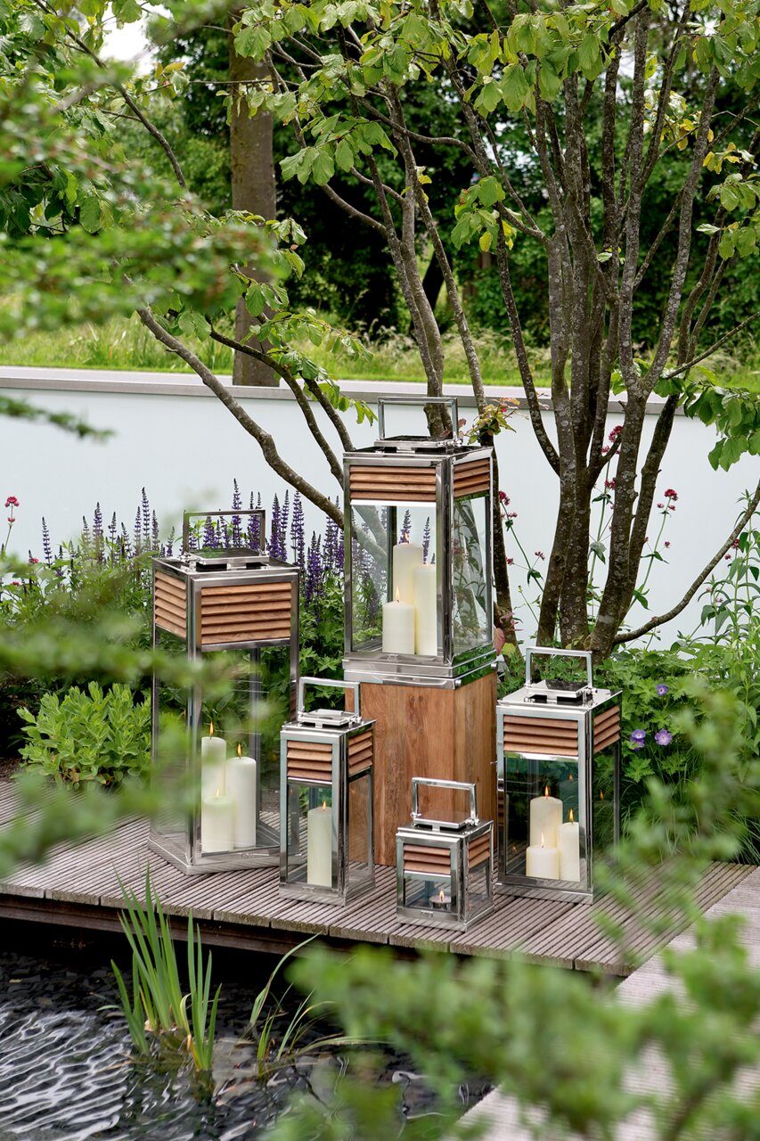 Verschillende lantaarns van glas en metaal met houten details, geplaatst op een terras in de tuin, omringd door groene planten. Aanzicht van voren.