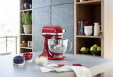 Rote Küchenmaschine mit Edelstahlschüssel, seitliche Perspektive, auf einer Küchenarbeitsplatte mit frischen Beeren und Sahne.
