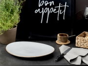Weißer, flacher Speiseteller mit feiner Sprenkelglasur und braunem Rand auf dunkler Tischplatte, im Vordergrund aus leicht schräger Seitenperspektive fotografiert; im Hintergrund Küchen-Accessoires wie Kräutertopf, Holzschale und Serviette mit Messer.