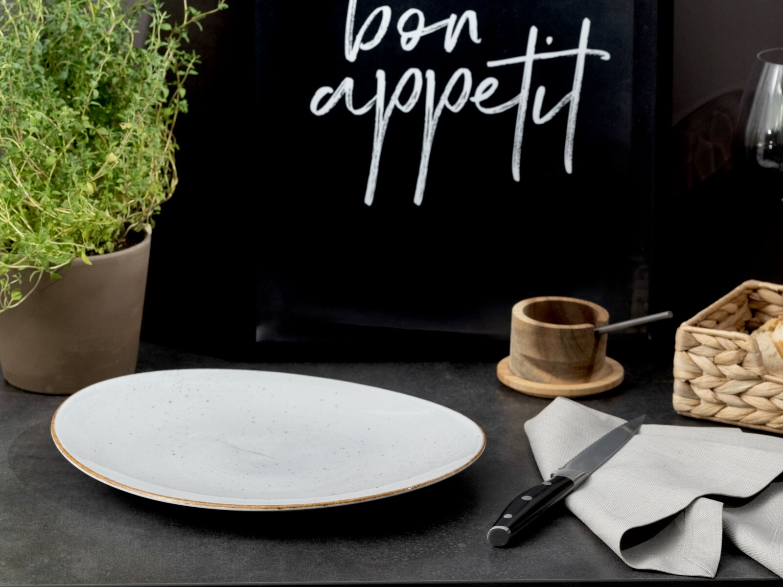 Weißer, flacher Speiseteller mit feiner Sprenkelglasur und braunem Rand auf dunkler Tischplatte, im Vordergrund aus leicht schräger Seitenperspektive fotografiert; im Hintergrund Küchen-Accessoires wie Kräutertopf, Holzschale und Serviette mit Messer.