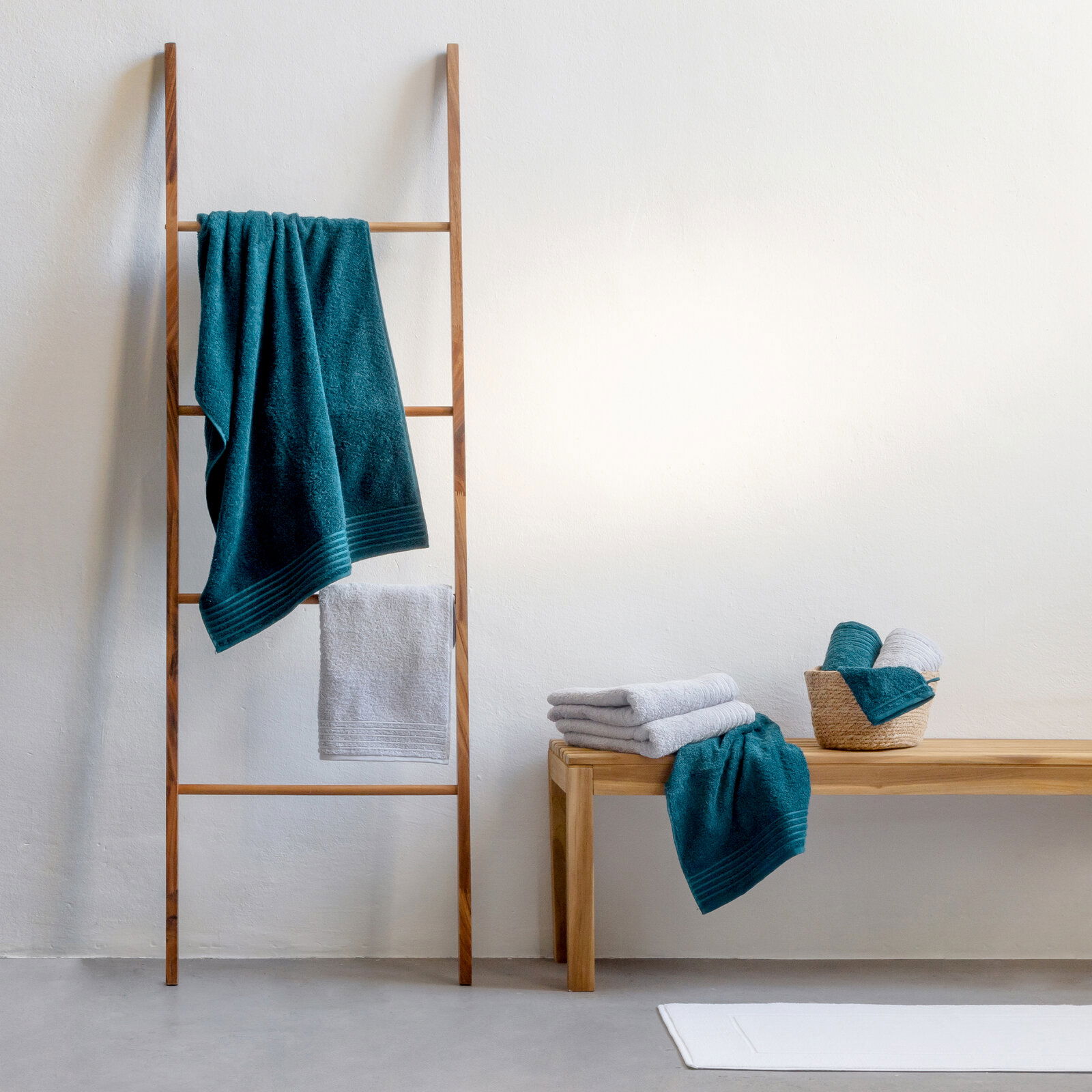 Handtuchset New Classic 10-tlg in einem modernen Badezimmer, mit einem Holzleiter-Handtuchhalter und einer Holzbank, auf der Handtücher gestapelt und in einem Korb arrangiert sind. Perspektive von vorne.