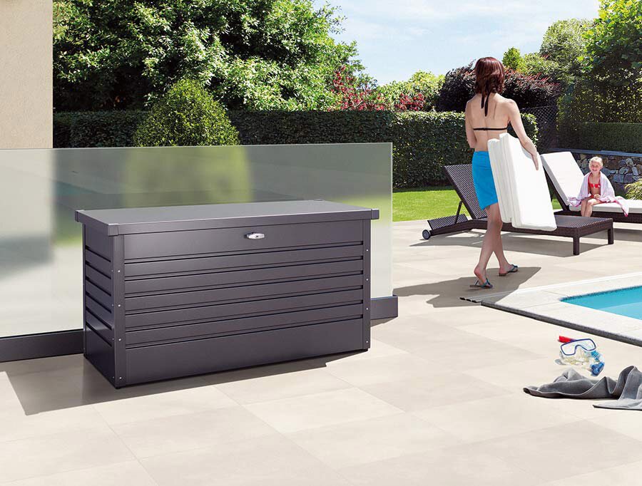 Dunkelgraue Garten-Aufbewahrungsbox aus Metall mit Deckel und Schloss vor einer Glaswand auf einer Poolterrasse, Perspektive: frontale leicht seitliche Ansicht