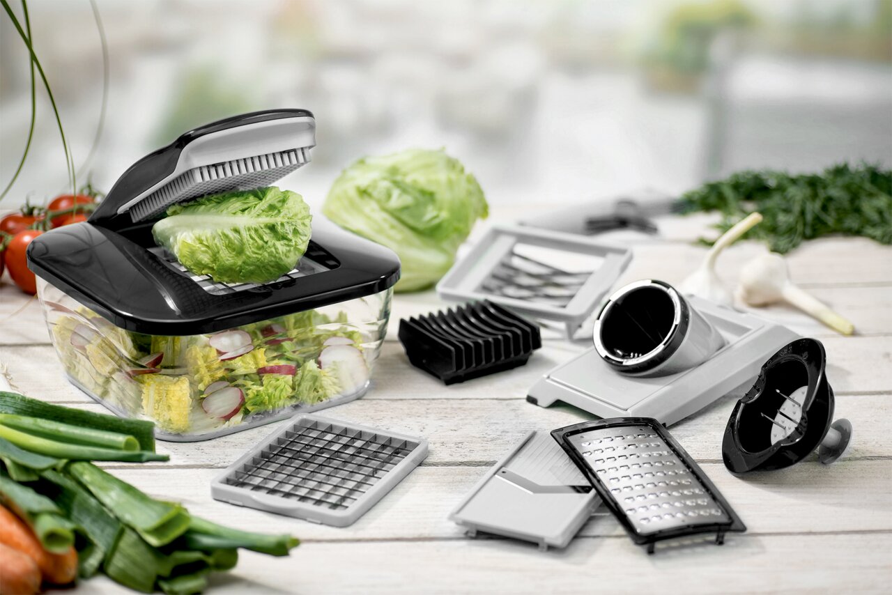 Multifunktionaler Gemüseschneider mit Auffangbehälter und verschiedenen Einsätzen (Hobel, Reibe, Würfelschneider) auf Holztisch, gefüllt mit Salat und Radieschenscheiben Perspektive: leicht schräg von vorne in Nahaufnahme.