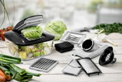 Multifunktionaler Gemüseschneider mit Auffangbehälter und verschiedenen Einsätzen (Hobel, Reibe, Würfelschneider) auf Holztisch, gefüllt mit Salat und Radieschenscheiben; Perspektive: leicht schräg von vorne in Nahaufnahme.