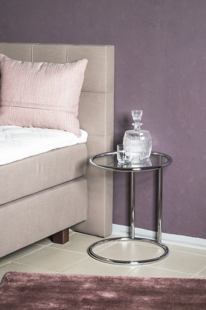 Runder Beistelltisch mit verchromtem Metallgestell und Glasplatte neben einem gepolsterten Bett, mit Glas-Karaffe und Trinkglas darauf, Ansicht schräg von vorn auf Augenhöhe.
