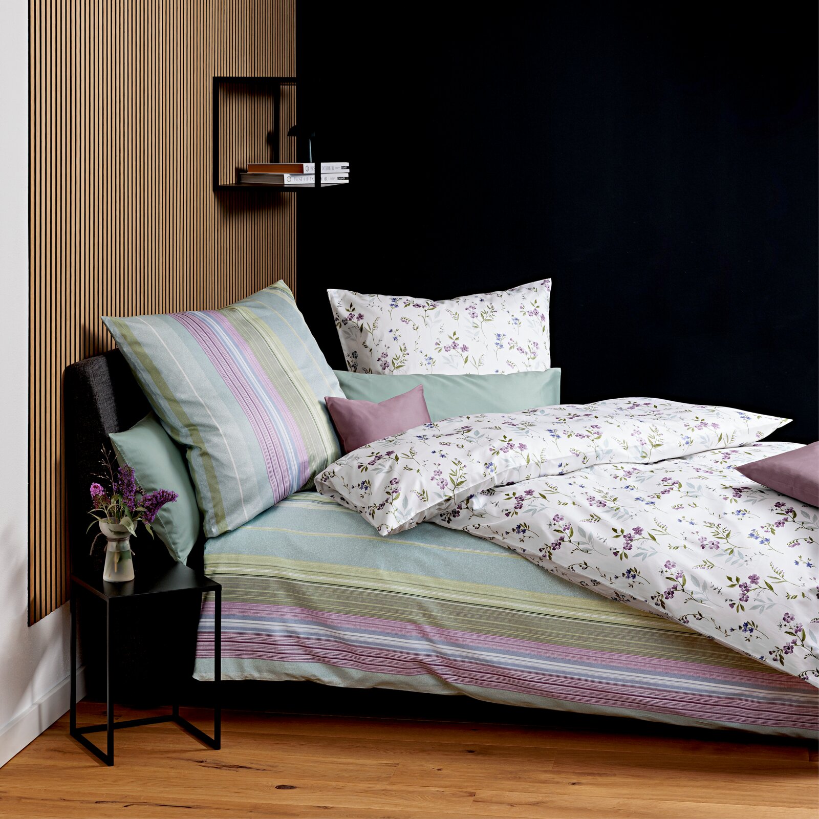 Schlafzimmer-Setting mit Bettwäsche: weißer Bettbezug mit zartem Blumenmuster, mintgrünen Kissen und gestreiftem Spannbettlaken in Pastelltönen; perspektivisch schräg von vorne links aufgenommen.