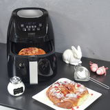 Schwarze Heißluftfritteuse mit digitalem Display, geöffnet mit Gebäck im Korb, auf einer Küchenarbeitsplatte von vorne fotografiert. Daneben ein Teller mit Gebäck, ein Puderzuckerstreuer und eine weiße Hasenfigur.