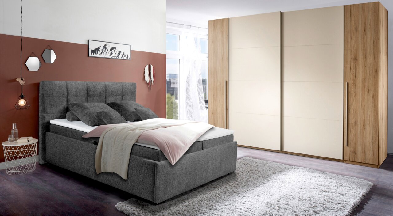 Graues Polsterbett mit hohem Kopfteil, seitlicher Blick, dekoriert mit Kissen und Decke, in modernem Schlafzimmer mit Teppich und Kleiderschrank.