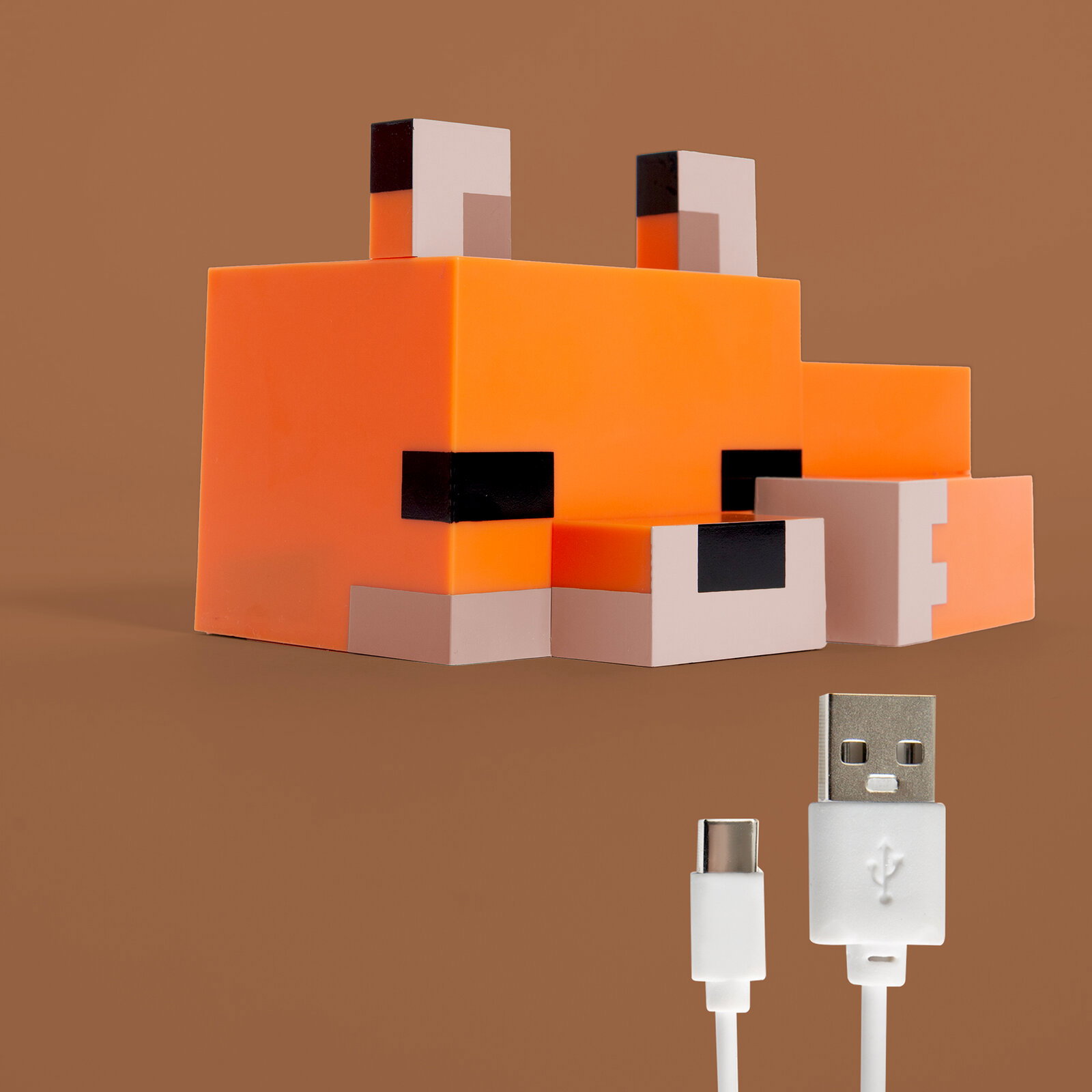Orangefarbene, würfelförmige Fuchs-Lampe im Pixel-/Block-Design mit schwarzen Augen und weißen Details, schräg von vorn links fotografiert; daneben ein weißes USB-A-auf-USB-C-Ladekabel vor braunem Hintergrund.