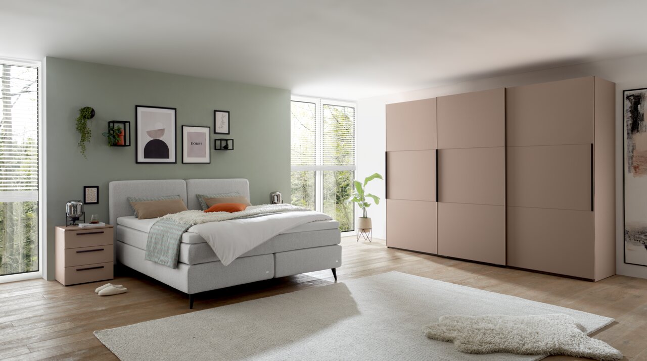 Modernes Boxspringbett in einem stilvoll eingerichteten Schlafzimmer mit grüner Wand, dekorativen Bildern und einem großen Kleiderschrank. Perspektive von der Seite.