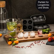 Runde Eiswürfelform mit fertigen Eiskugeln auf einem Holztablett, umgeben von Getränken und Früchten, aus der Perspektive von vorne.