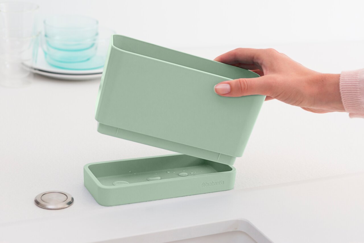 Brabantia Spül-Organizer SINKSIDE Grüner Spülen Organizer in der Hand gehalten, mit abnehmbarem Boden, auf einer weißen Arbeitsfläche.