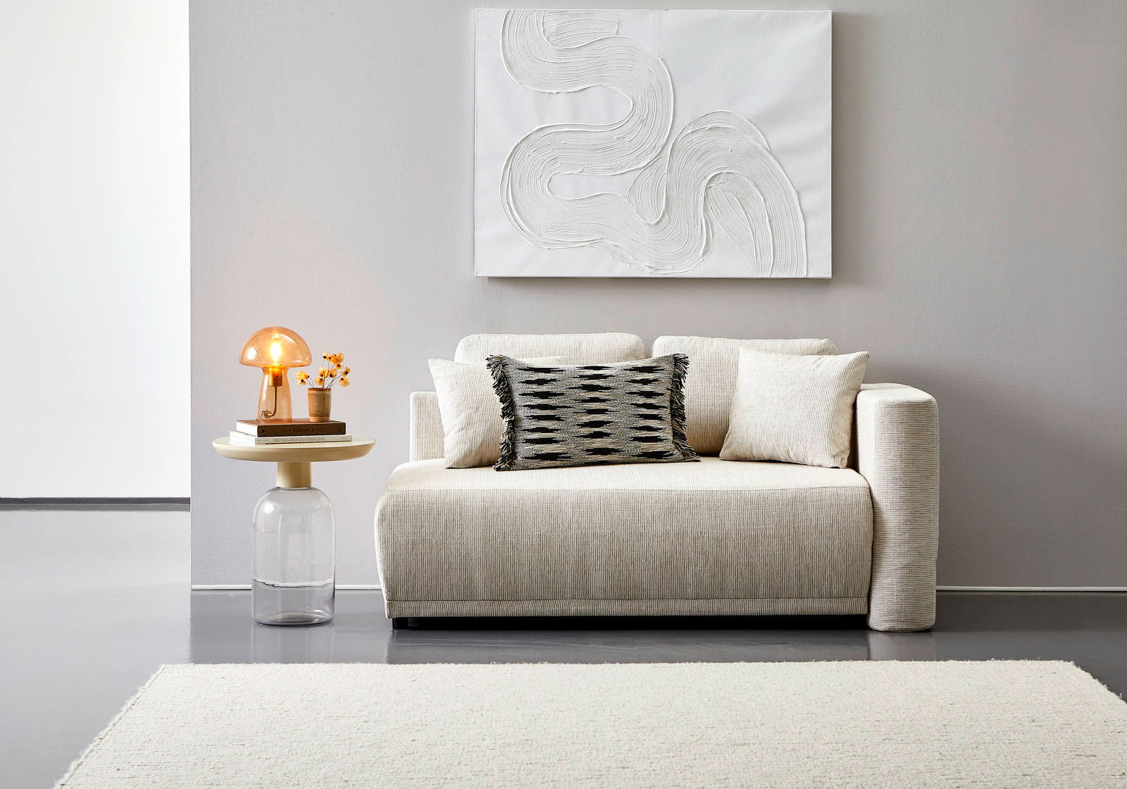 Frontale Ansicht einer eleganten, cremefarbenen Recamiere mit dekorativen Kissen, neben einem modernen Beistelltisch mit Lampe und Blumen.