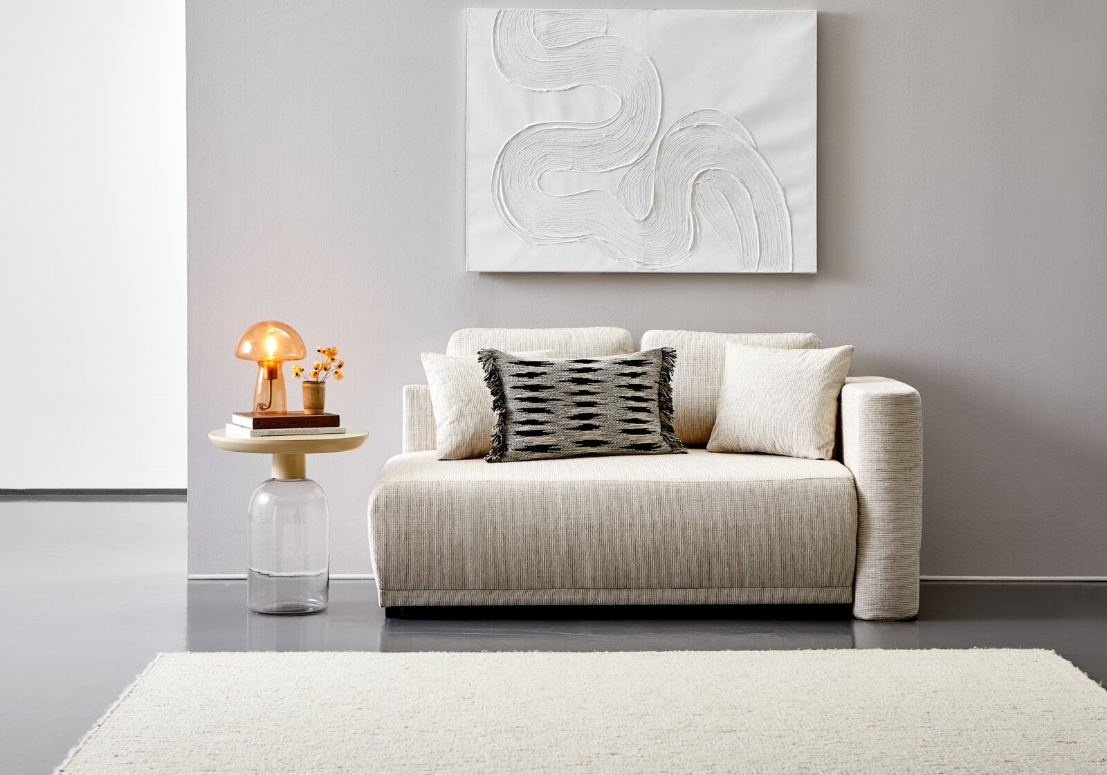 Vooraanzicht van een elegante, crèmekleurige recamier met decoratieve kussens, naast een modern bijzettafeltje met een lamp en bloemen.