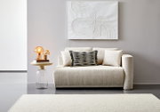 Vooraanzicht van een elegante, crèmekleurige recamier met decoratieve kussens, naast een modern bijzettafeltje met een lamp en bloemen.