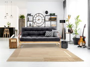 Moderner Wohnraum mit einem beigen Wollteppich im Vordergrund, der vor einem grauen Sofa liegt. Der Raum ist stilvoll eingerichtet mit Pflanzen, einer Gitarre und einem großen Wanduhr. Die Perspektive zeigt den Teppich von vorne.