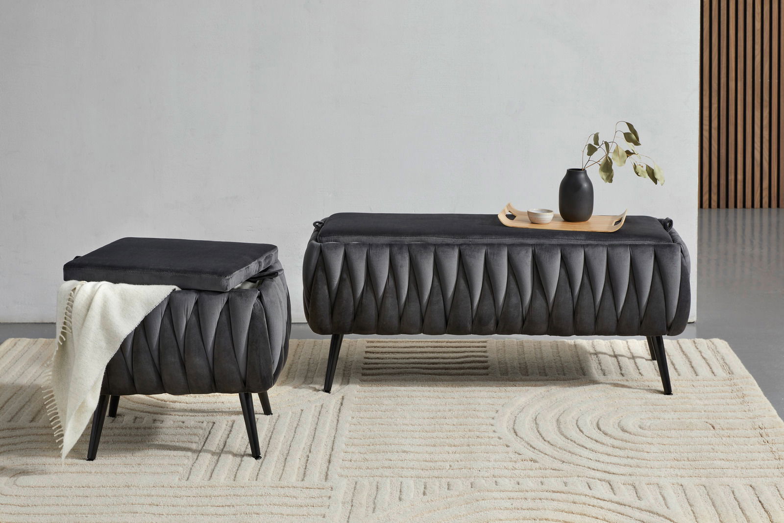 Zwei moderne, dunkelgraue Hocker mit eleganter, gefalteter Polsterung auf einem hellen Teppich. Einer der Hocker ist mit einer weißen Decke dekoriert, während der andere ein Tablett mit einer Vase und Schalen trägt. Perspektive: Frontalansicht.