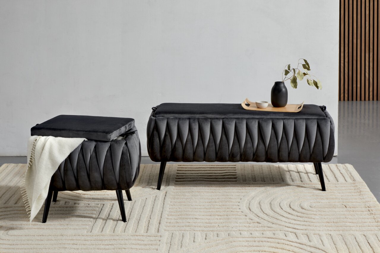 Zwei moderne, dunkelgraue Hocker mit eleganter, gefalteter Polsterung auf einem hellen Teppich. Einer der Hocker ist mit einer weißen Decke dekoriert, während der andere ein Tablett mit einer Vase und Schalen trägt. Perspektive: Frontalansicht.