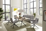 Schuin vooraanzicht van een moderne eethoek met een licht houten tafel, grijze draaistoelen en een bijpassende hoekbank in een lichte ruimte met hoge ramen.