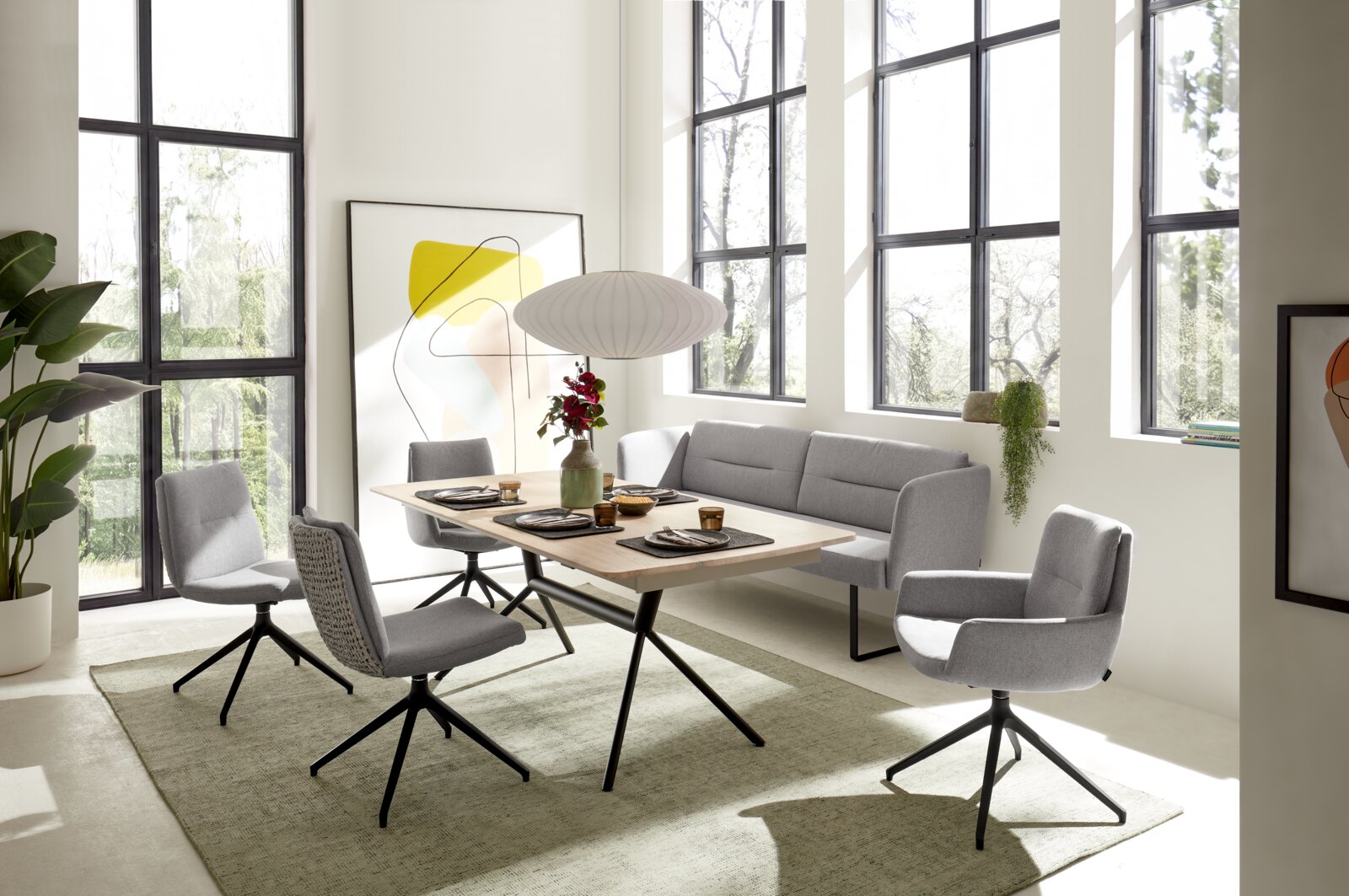Schuin vooraanzicht van een moderne eethoek met een licht houten tafel, grijze draaistoelen en een bijpassende hoekbank in een lichte ruimte met hoge ramen.