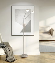 Moderne LED-Stehlampe in Beige mit zwei verstellbaren Leuchtenköpfen und rundem Standfuß, frontal in einem hellen Wohnzimmer neben Sofa und Wandbild aufgenommen.