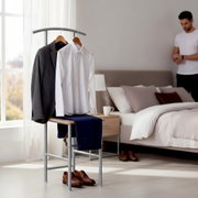 Moderner Herrendiener/Kleiderständer aus Metall im Schlafzimmer, mit grauem Sakko und weißem Hemd auf Bügeln sowie blauer Hose über der Ablage; braune Lederschuhe davor, aufgenommen in frontaler Perspektive leicht von links.