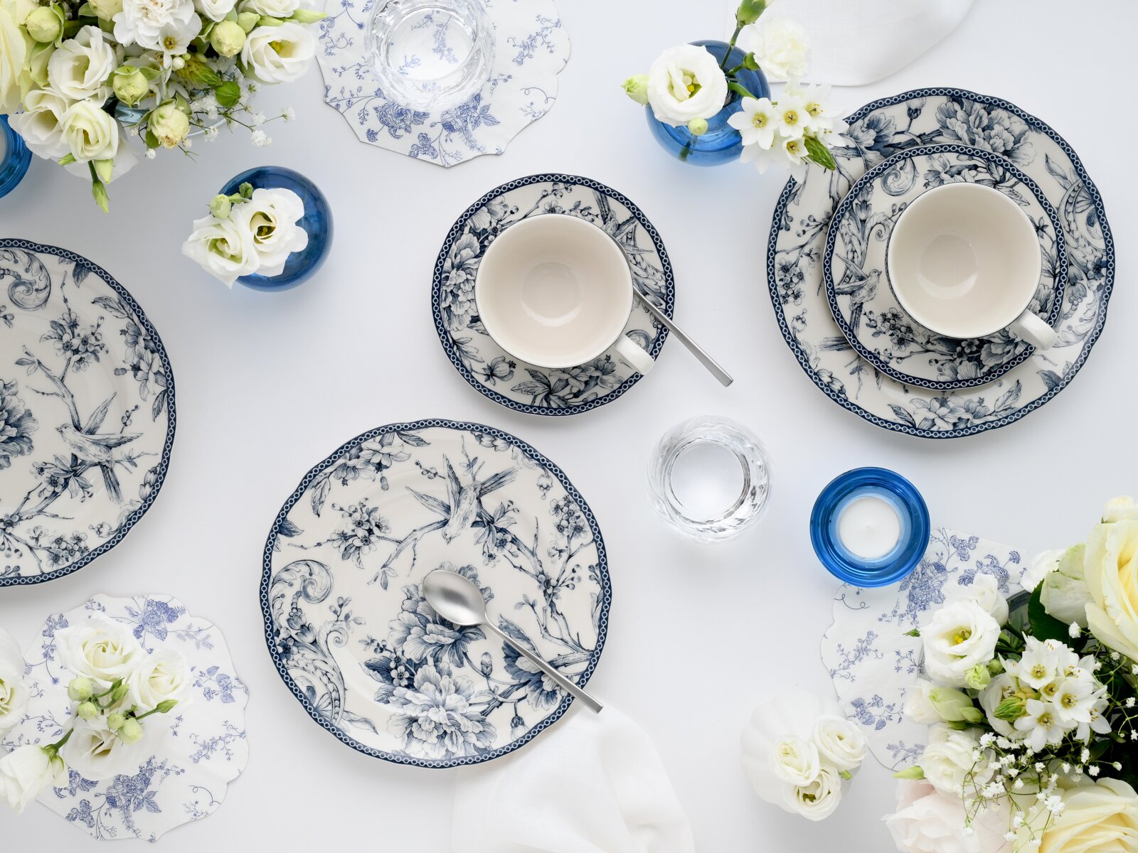 Draufsicht (Flatlay) auf ein blau-weißes Porzellan-Tafelservice mit floralen Mustern: Teller, Tassen mit Untertassen, Löffel, Glas und blaue Vasen mit weißen Blumen auf hellem Tisch