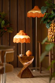 Retro Stehleuchte mit orangefarbenem Fransen-Lampenschirm, im Vordergrund aus seitlicher Perspektive fotografiert, umgeben von Holzmöbeln und Pflanzen.