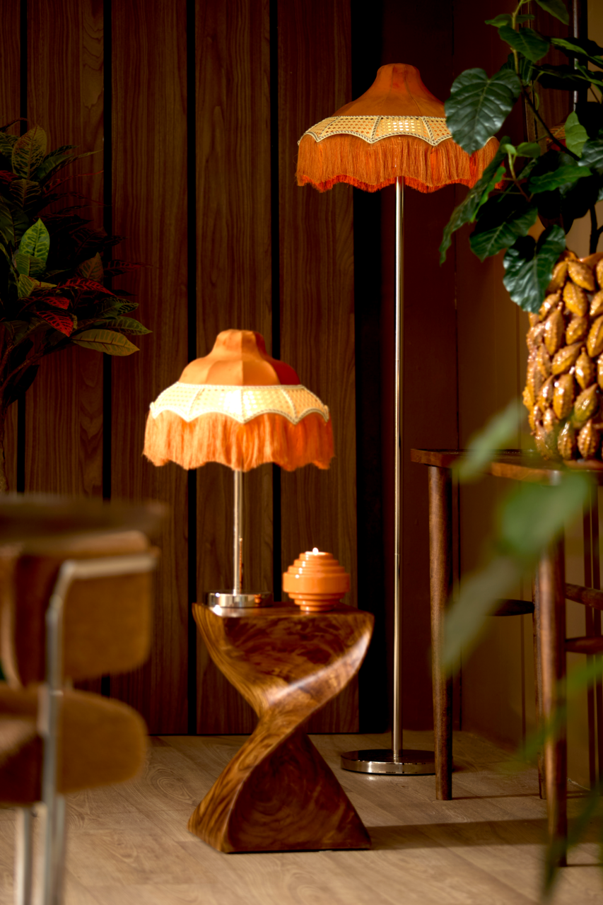 Retro Stehleuchte mit orangefarbenem Fransen-Lampenschirm, im Vordergrund aus seitlicher Perspektive fotografiert, umgeben von Holzmöbeln und Pflanzen.