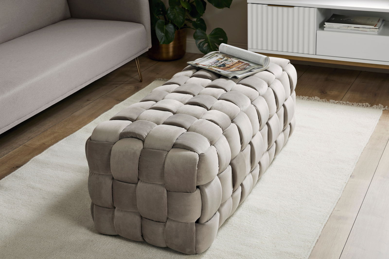 Beiger, gepolsterter Hocker in rechteckiger Form mit geflochtenem Design, auf einem Teppich in einem modernen Wohnzimmer, seitliche Perspektive.