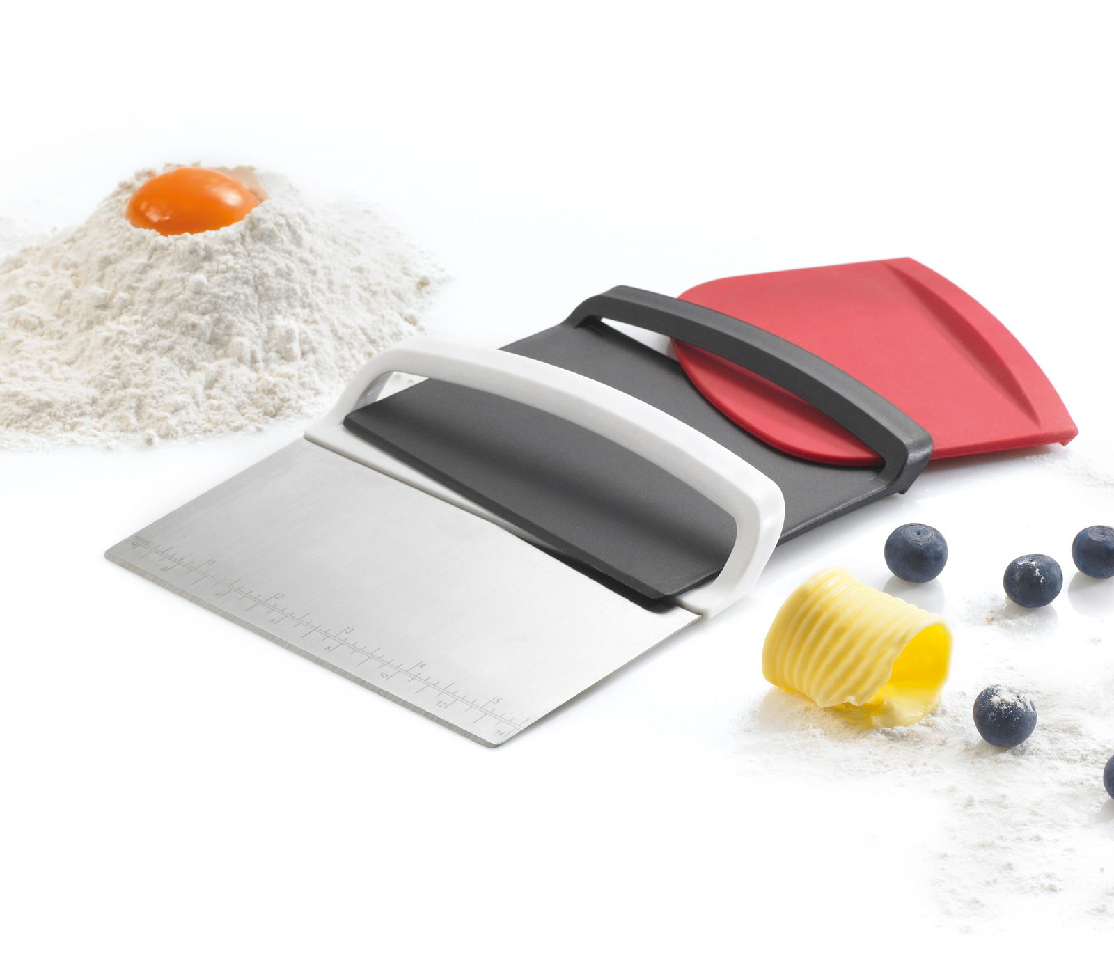 Teigschaber-Set 3-tlg. in seitlicher Perspektive mit Edelstahlklinge und zwei Kunststoffschabern in Schwarz und Rot, umgeben von Mehl, Ei, Butter und Blaubeeren.