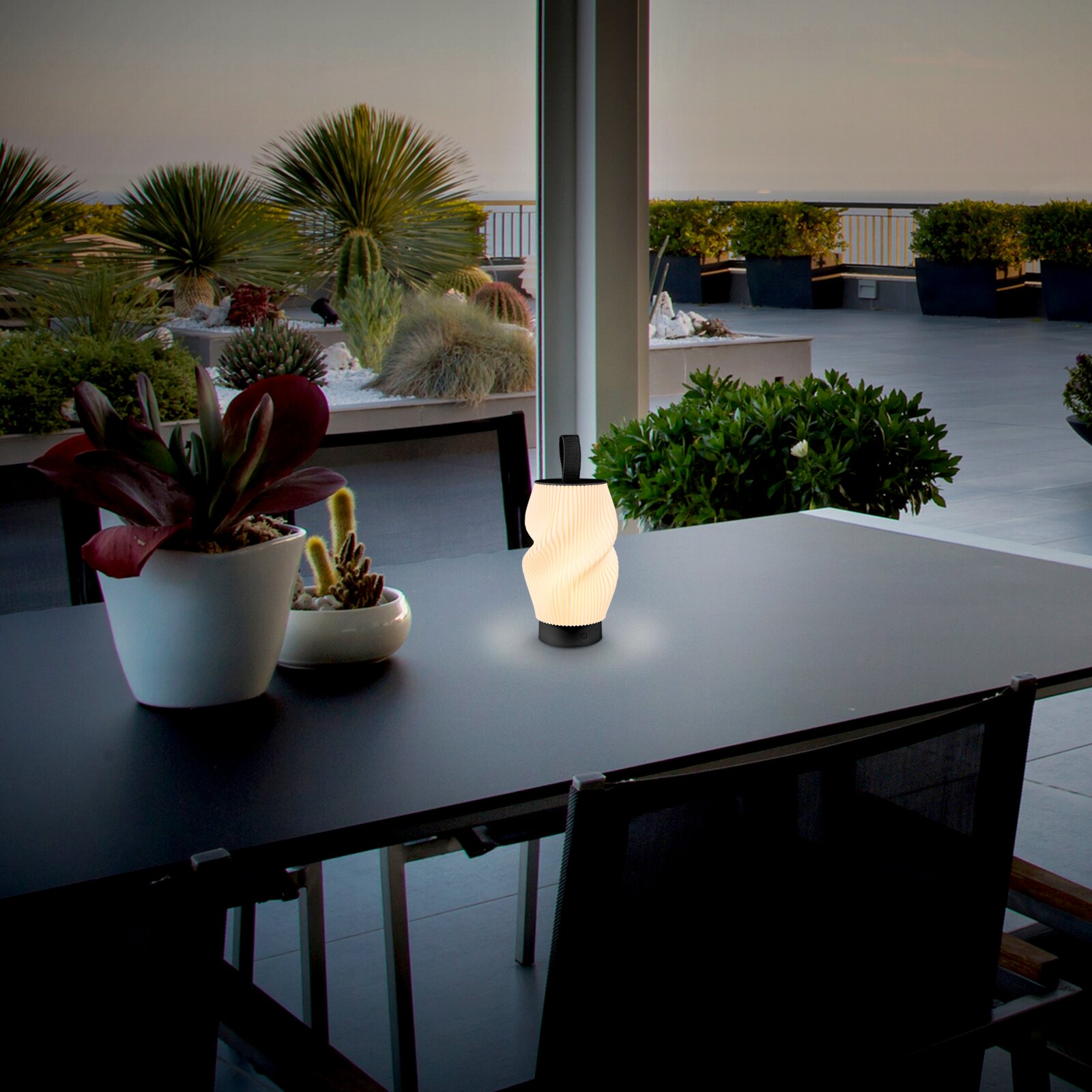Leuchtende, tragbare LED-Tischlampe mit gewelltem Diffusor und schwarzem Sockel auf einem dunklen Outdoor-Tisch auf einer Dachterrasse, Perspektive: leicht seitlich von vorne auf Tischhöhe.