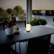 Leuchtende, tragbare LED-Tischlampe mit gewelltem Diffusor und schwarzem Sockel auf einem dunklen Outdoor-Tisch auf einer Dachterrasse, Perspektive: leicht seitlich von vorne auf Tischhöhe.