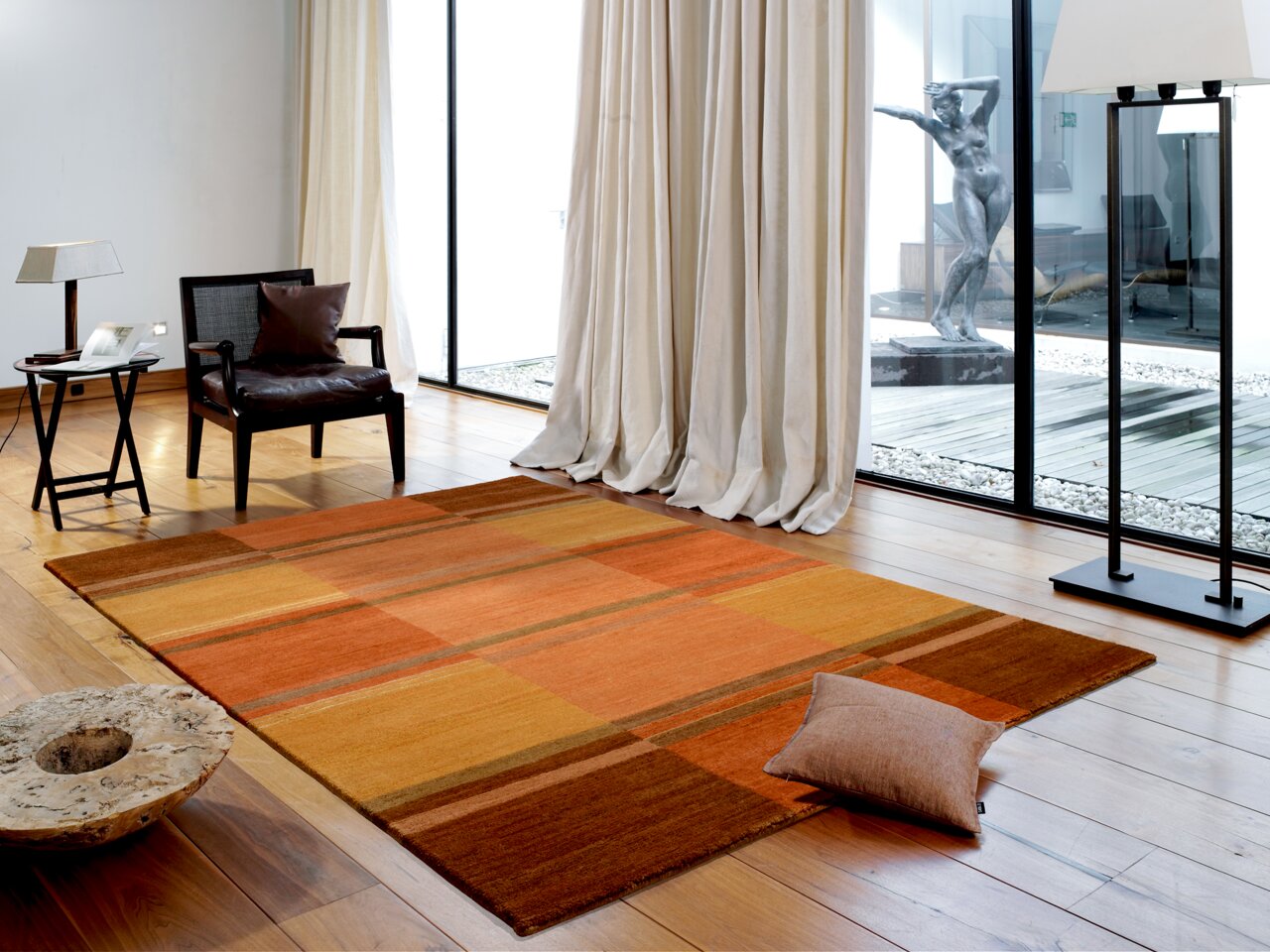 Wollteppich in warmen Orangetönen in einem modernen Wohnzimmer mit Holzboden, einem Sessel und großen Fenstern. Perspektive von oben.