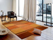 Wollteppich in warmen Orangetönen in einem modernen Wohnzimmer mit Holzboden, einem Sessel und großen Fenstern. Perspektive von oben.