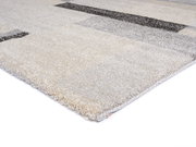 Nahaufnahme eines rechteckigen Teppichs mit geometrischem Muster in Beige- und Grautönen, perspektivisch von der Seite aufgenommen.