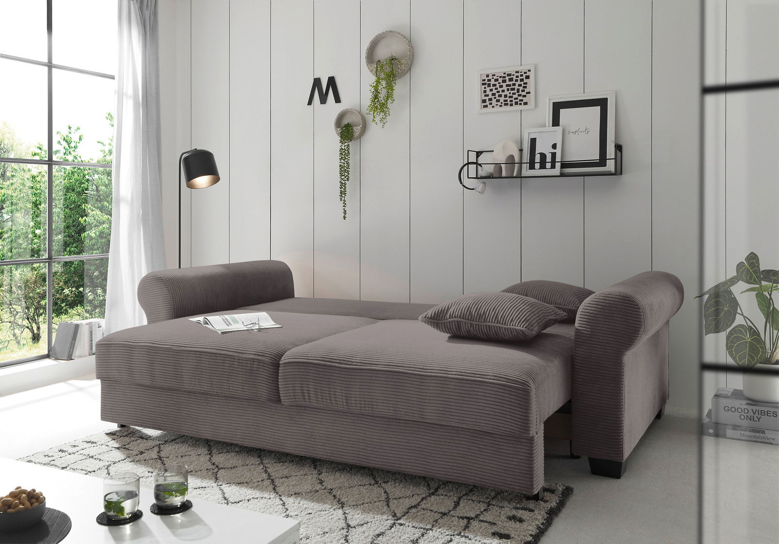 Graues Funktionssofa in einem modernen Wohnzimmer, seitliche Perspektive, mit dekorativen Kissen und Pflanzen im Hintergrund.