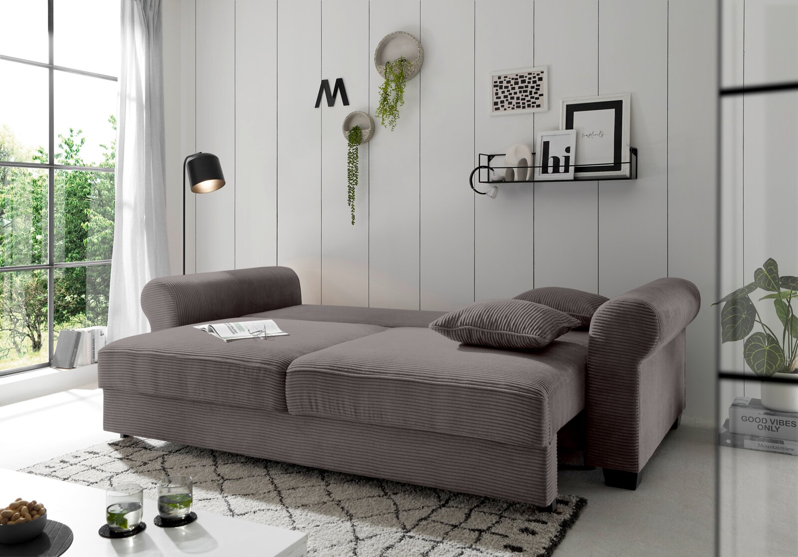 Grijze functionele bank in een moderne woonkamer, gezien vanaf de zijkant, met sierkussens en planten op de achtergrond.