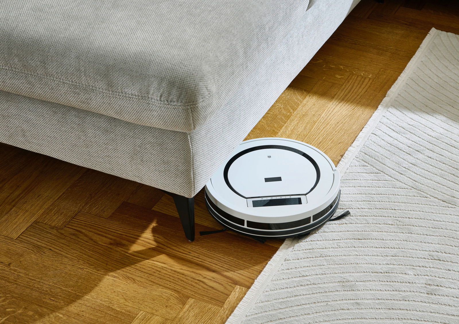 Ein weißer Saugroboter reinigt den Boden neben einem grauen Sofa aus der Vogelperspektive.