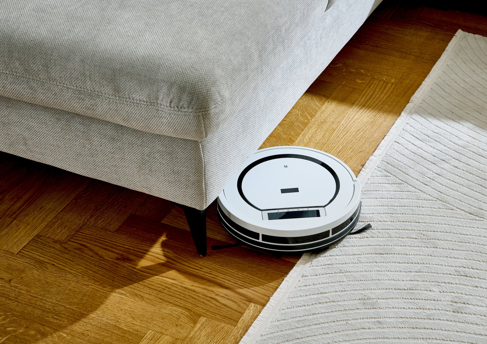 Ein weißer Saugroboter reinigt den Boden neben einem grauen Sofa aus der Vogelperspektive.