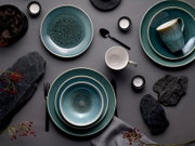 Draufsicht (Flatlay) auf ein blau-grün glasiertes Keramik-Geschirrset mit Tellern, Schalen und Tassen auf grauem Hintergrund, dekoriert mit schwarzen Besteckteilen, Teelichtern und dunklen Steinen.