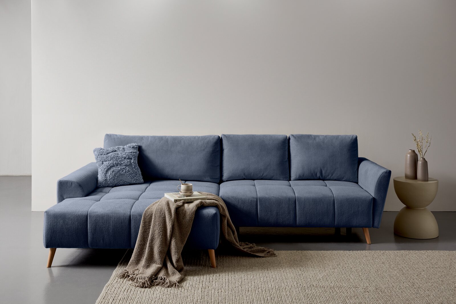 Z2 Ecksofa ZELDA Blaues, modern gepolstertes Ecksofa mit linksseitiger Ottomane, gesteppten Sitzflächen und Holzfüßen, frontal in leichter Schrägansicht fotografiert; davor Decke und Bücher, daneben Beistelltisch mit Vasen.