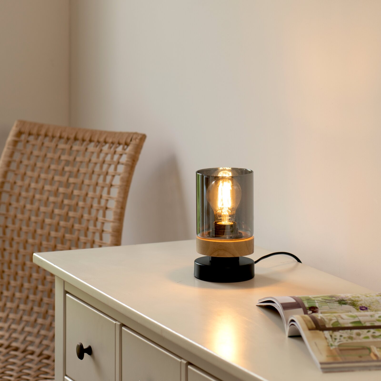 Just Light Tischleuchte PASQUAL Leuchtende Tischlampe mit rauchgrauem Glaszylinder, Holzring und schwarzem Sockel auf weißer Kommode neben aufgeschlagenem Magazin, Perspektive: leicht seitlich von vorne auf Tischhöhe