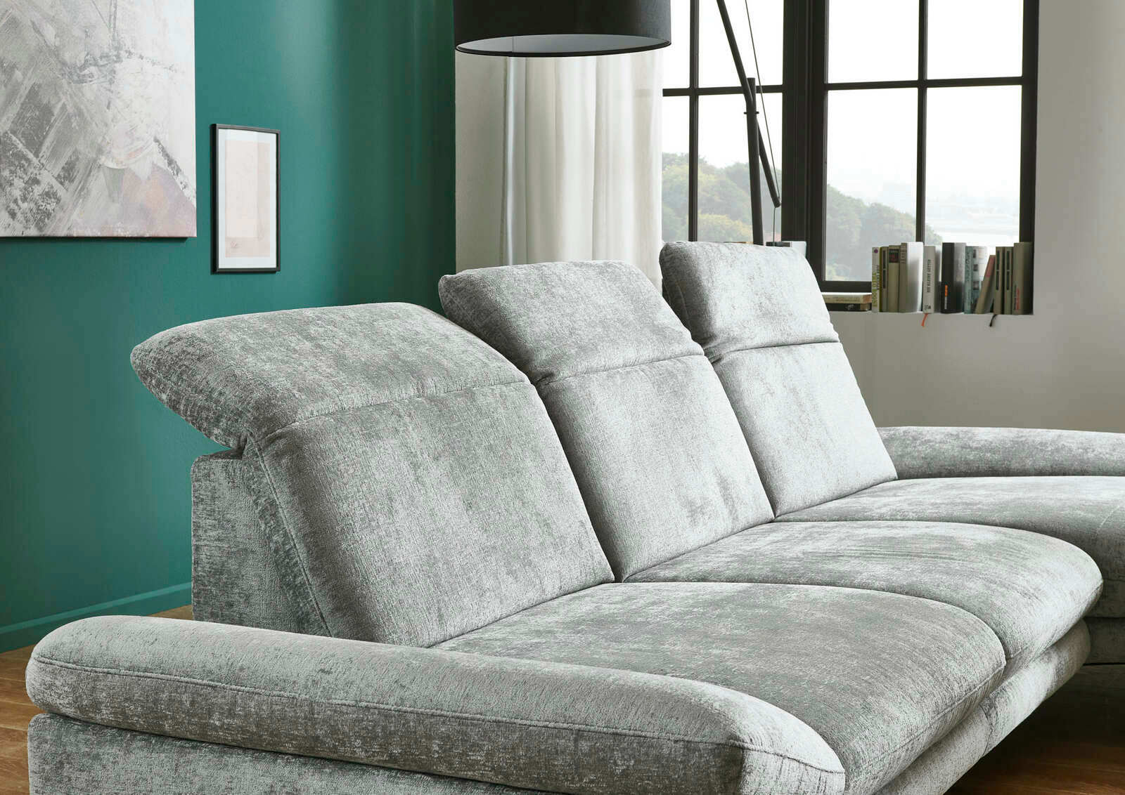 Graues Longchair-Sofa mit verstellbaren Kopfstützen, seitliche Perspektive in einem modernen Wohnzimmer mit grüner Wand und großen Fenstern.
