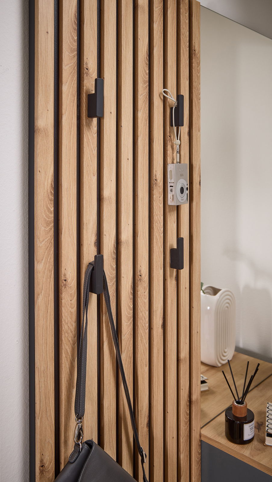 Nahaufnahme einer Kompaktgarderobe mit Holzlatten und schwarzen Haken, an denen eine Kamera und eine Tasche hängen. Perspektive von der Seite.