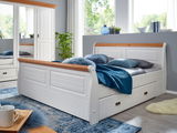 Weißes Holzbett mit Stauraum, seitliche Perspektive, dekoriert mit blauen Kissen und Decke, in einem hellen Raum mit großen Fenstern.